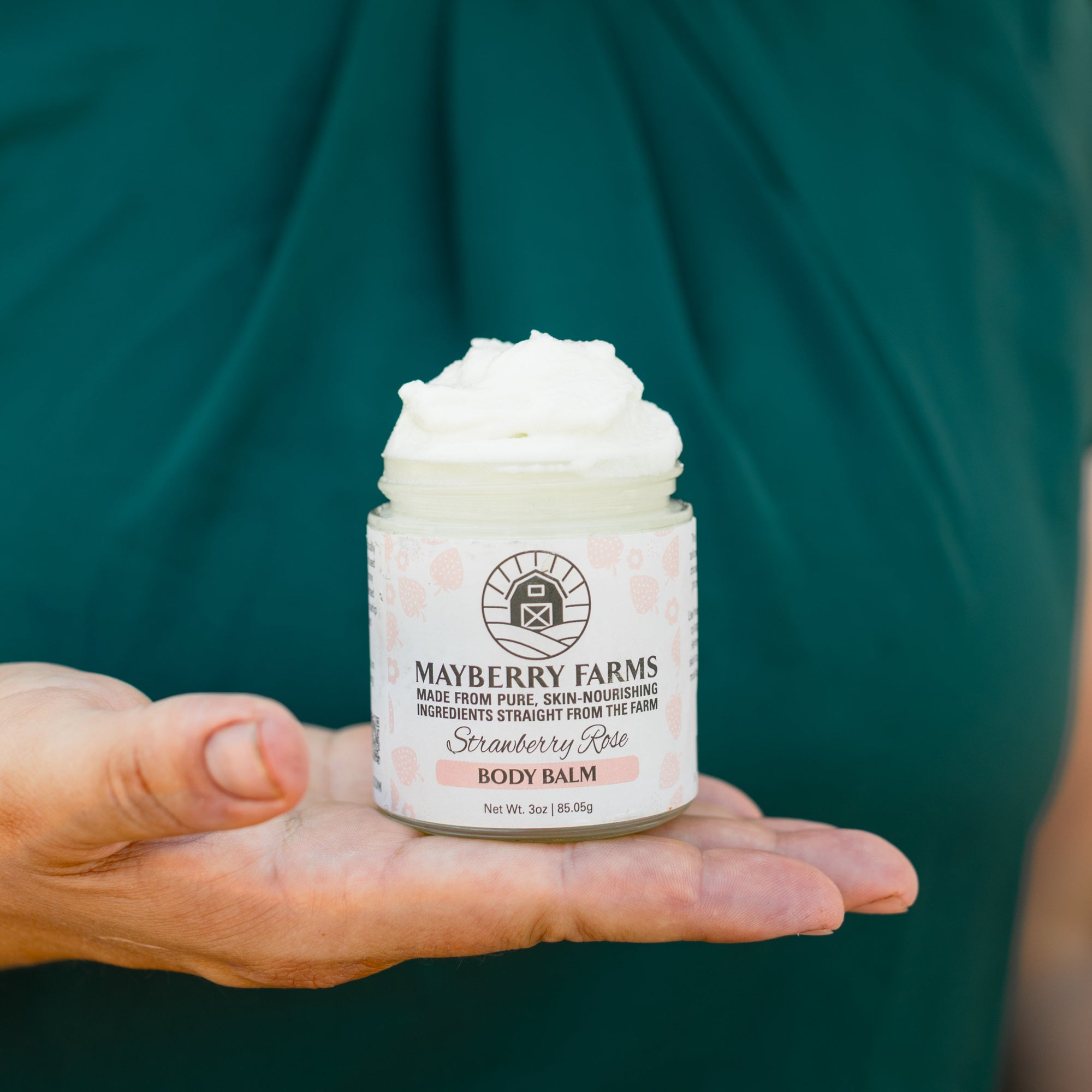 Person holding a jar of Mayberry Farms body balm against a green background