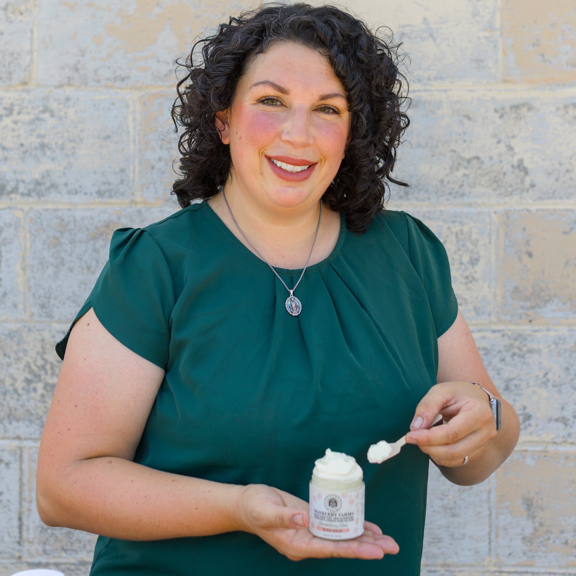 Woman holding a jar of cream and a applicator against a brick wall.