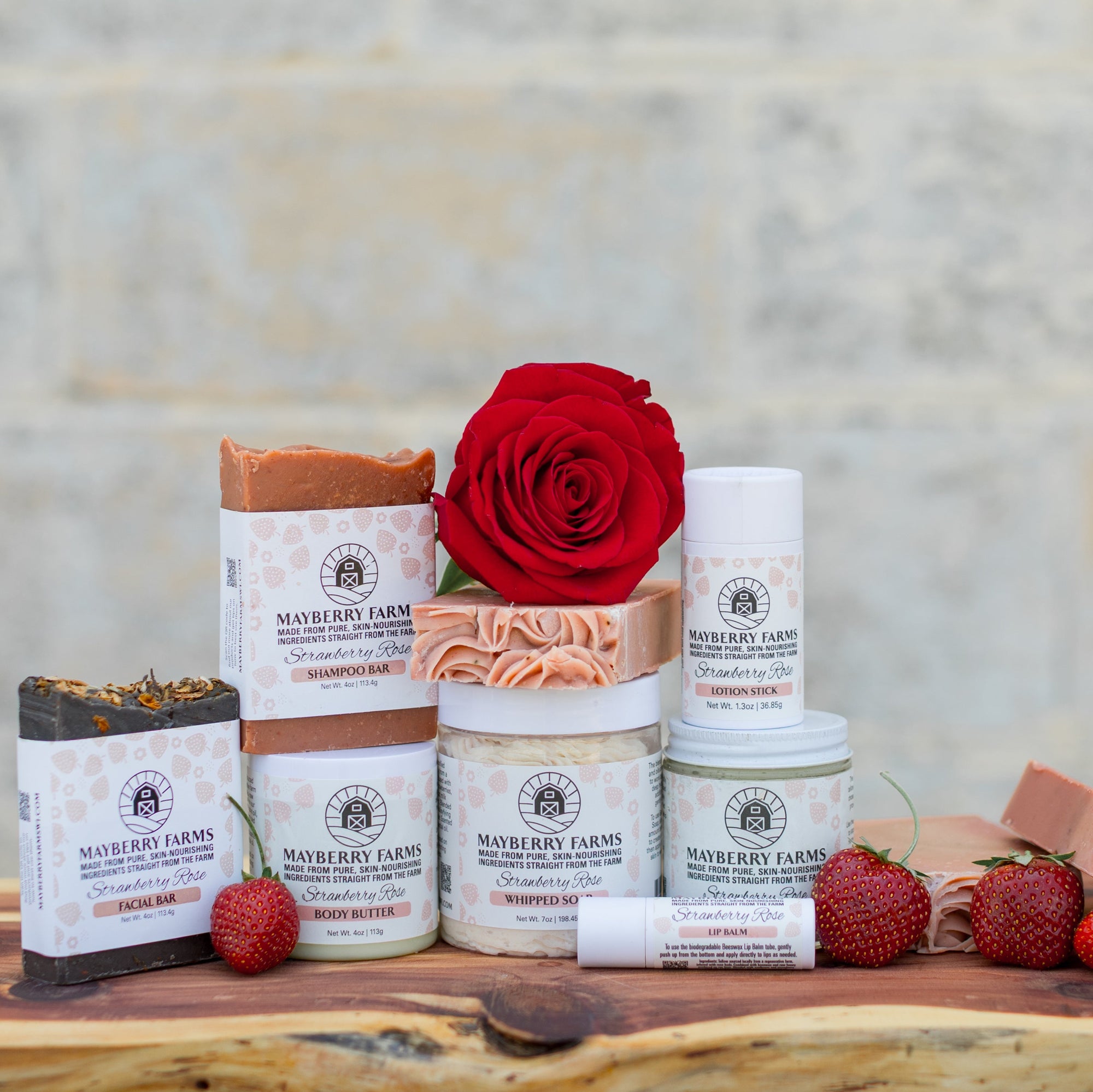Collection of Mad River Farm products including soaps, lip balm, and a rose on a wooden surface with a stone wall background.