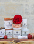 Collection of Mad River Farm products including soaps, lip balm, and a rose on a wooden surface with a stone wall background.